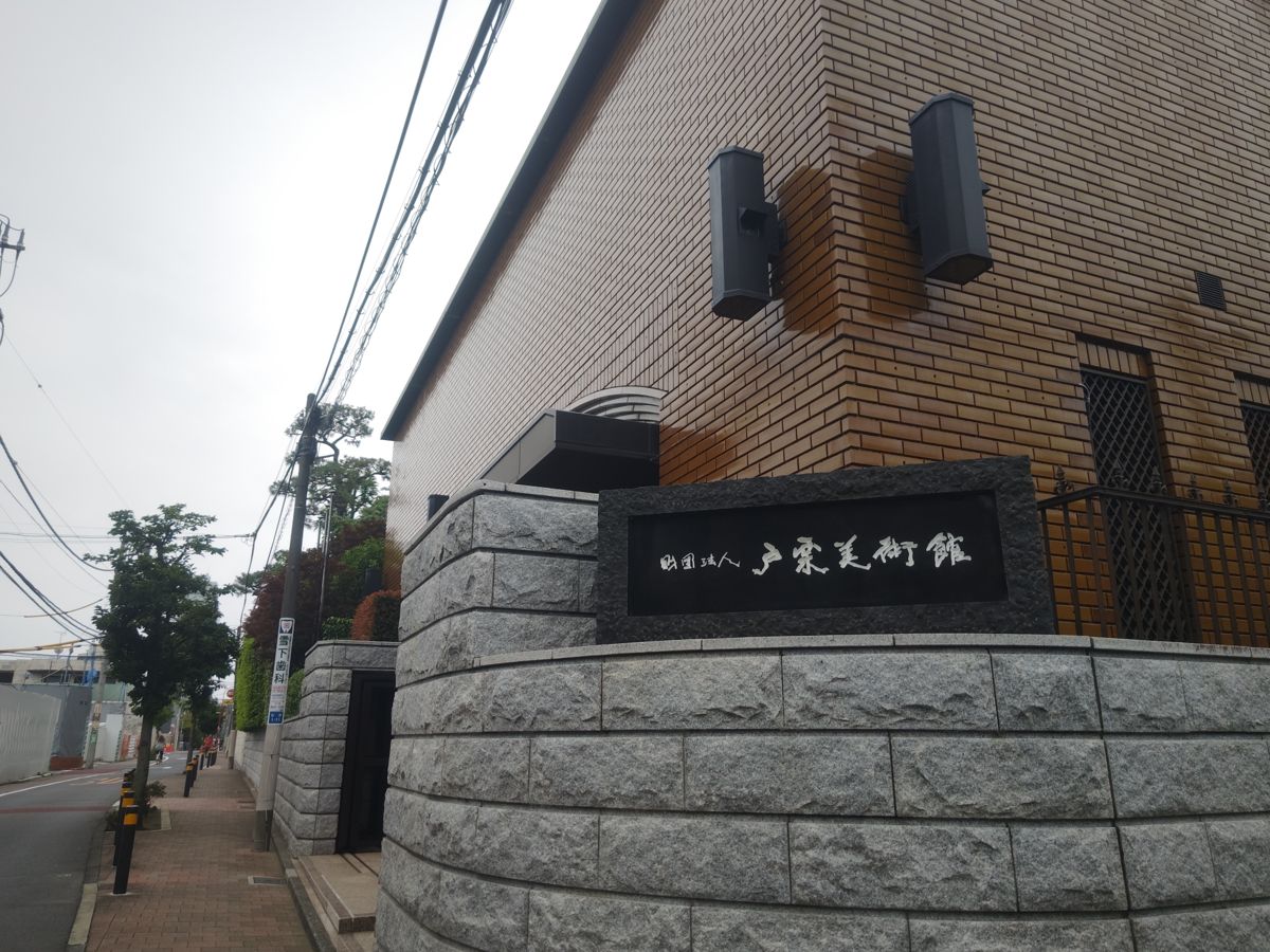 れんが色のタイル張りの建物の角に、石積みの台と「戸栗美術館」の銘板。歩道沿いに街路樹