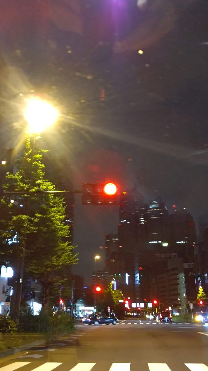 夜の交差点。街灯に照らされた街路樹の緑が浮かび、奥に高層ビルが並ぶ